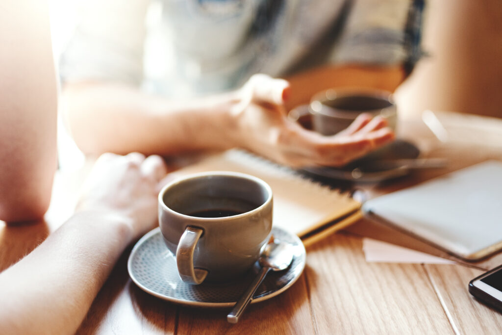 Friends talking at cafe table during coffee break. Unrecognizable male and female colleagues discussing business issues, focus on coffee cup with saucer and teaspoon